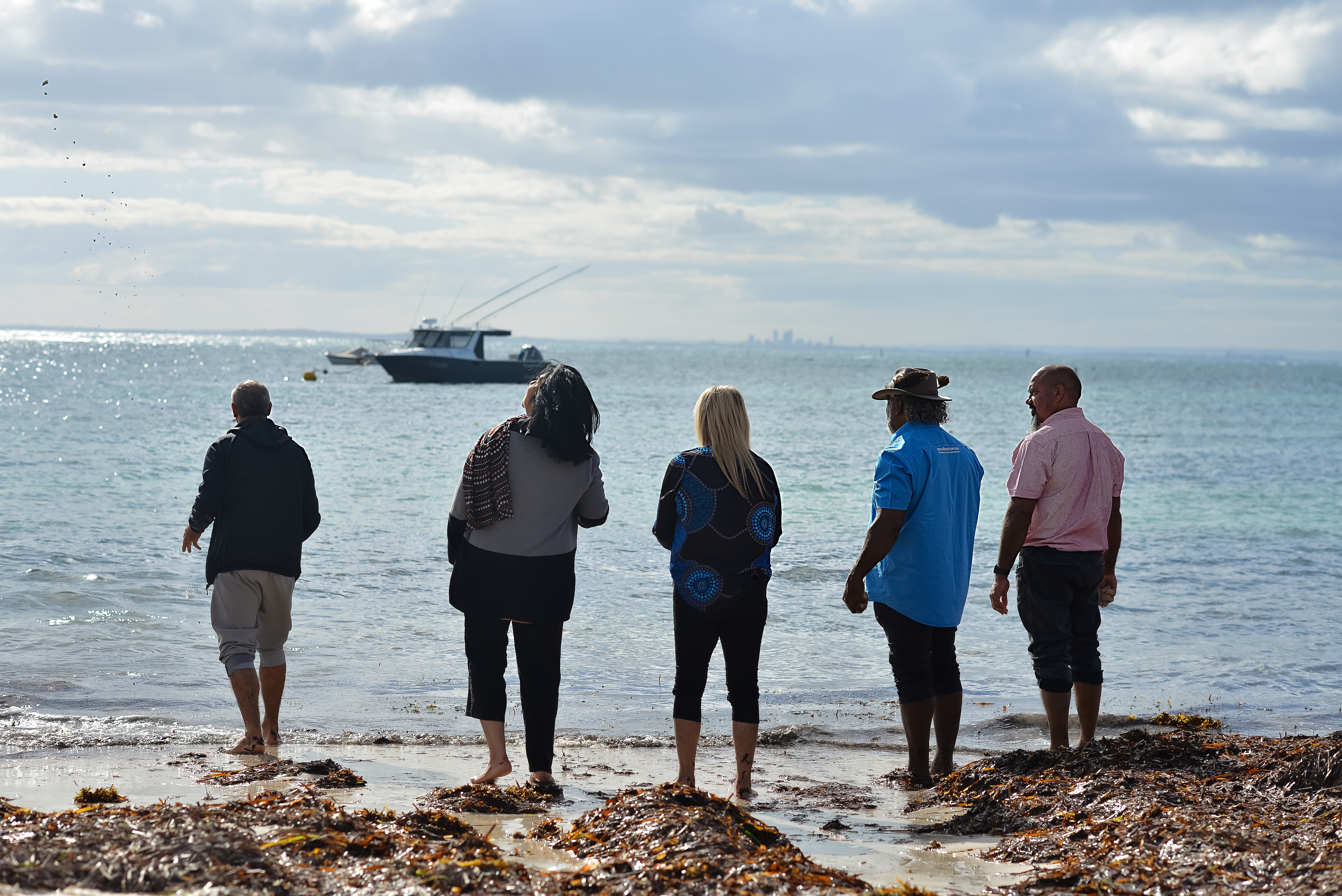 Five people standing at the shoreline, looking out to the ocean with a boat offshore.