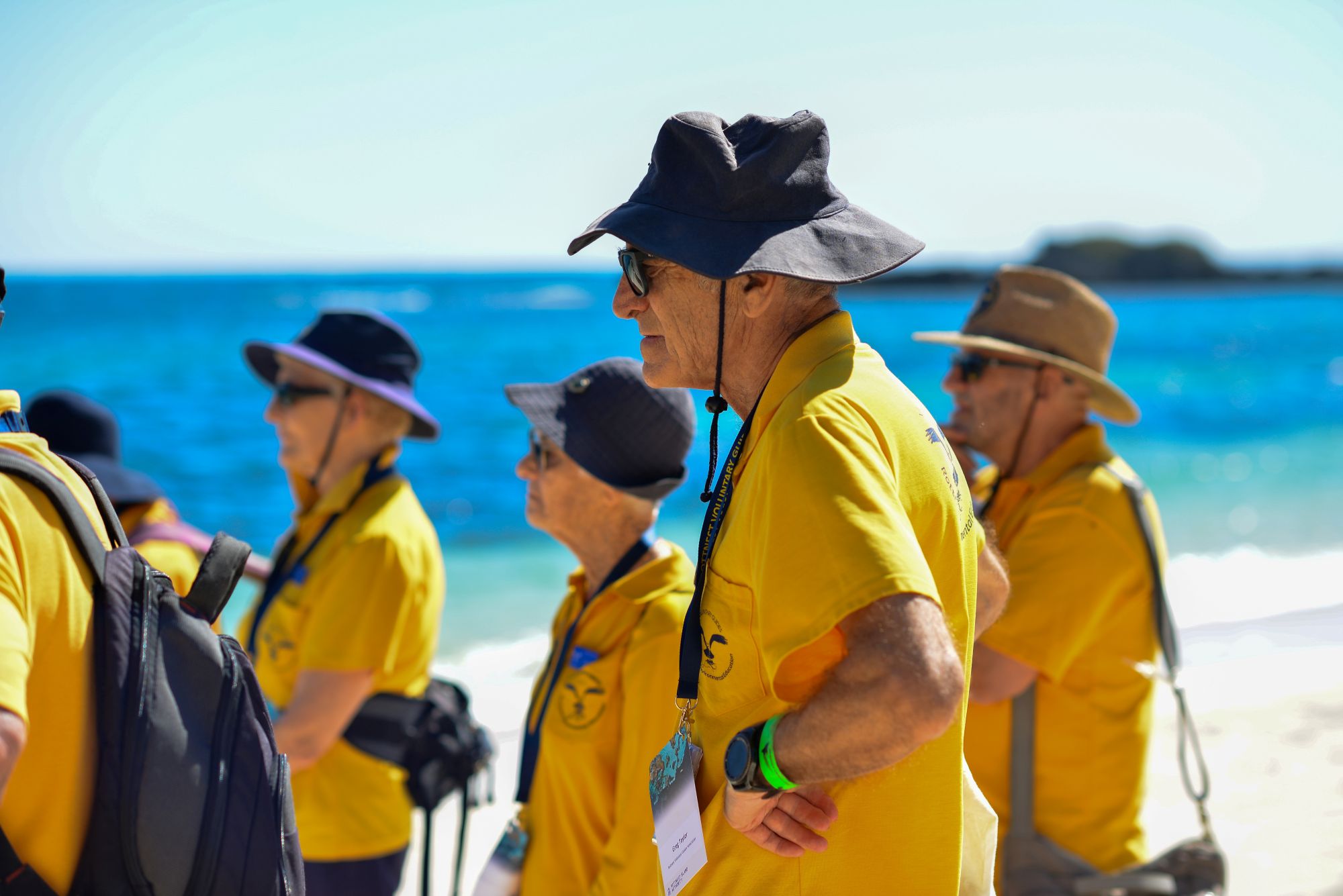 Volunteers of the Rottnest Voluntary Guides Association standing by the beach wearing hats and yellow polo shirts.